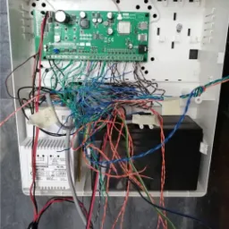 Wnętrze białej obudowy centrali alarmowej z widoczną płytą główną Satel Perfecta 16, akumulatorem i transformatorem AWT150, z licznymi przewodami o różnych kolorach podłączonymi do listwy zaciskowej.