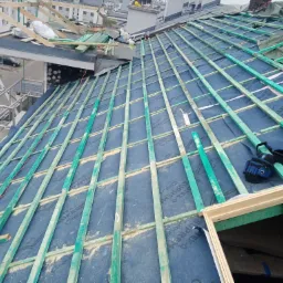 Konstrukcja dachu w trakcie budowy: widoczne drewniane łaty w kolorze zielonym, folia dachowa w kolorze niebieskim, fragment okna dachowego i narzędzia dekarskie leżące na dachu.