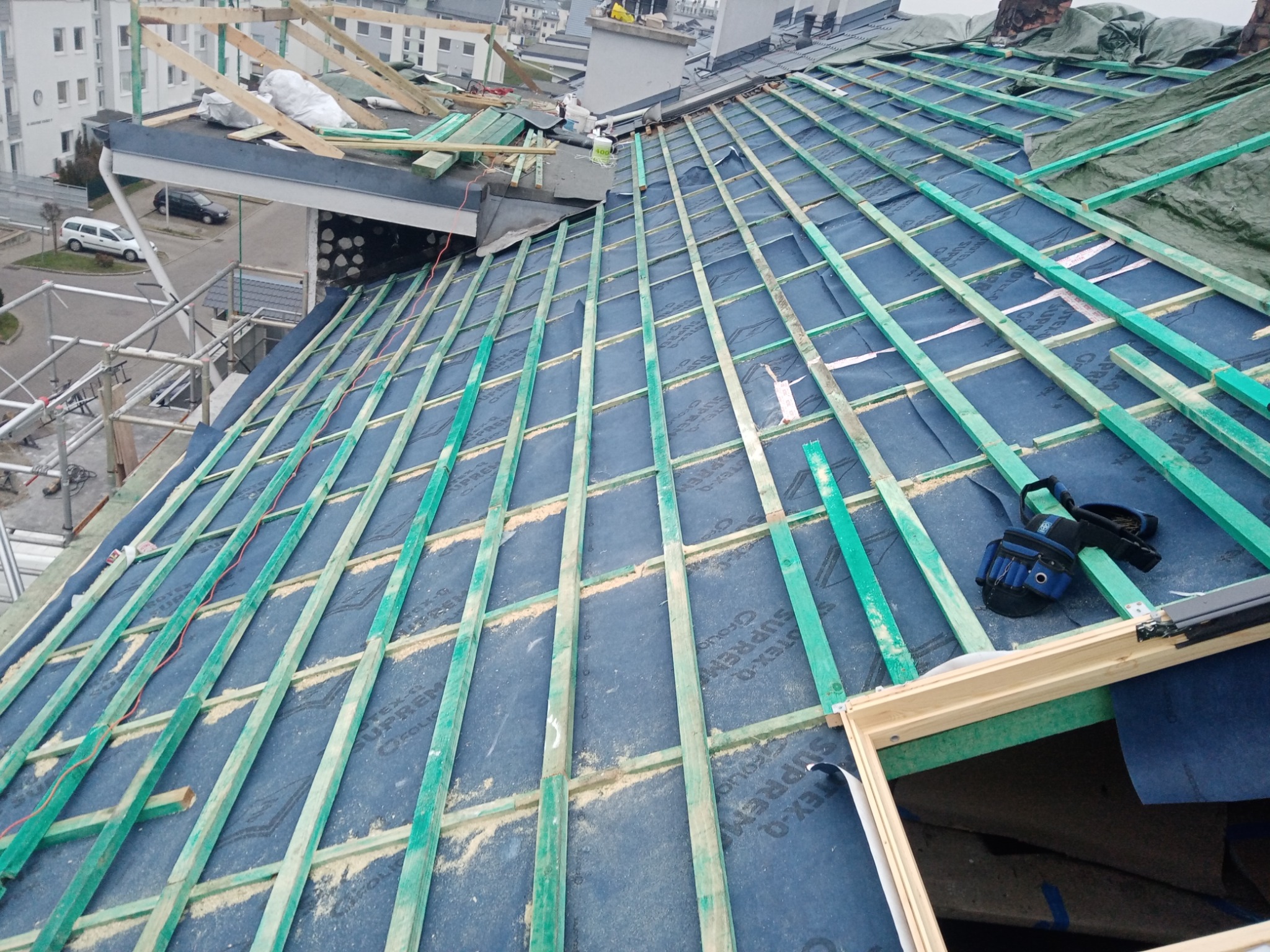 Konstrukcja dachu w trakcie budowy: widoczne drewniane łaty w kolorze zielonym, folia dachowa w kolorze niebieskim, fragment okna dachowego i narzędzia dekarskie leżące na dachu.