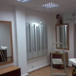 Wnętrze salonu optycznego w trakcie remontu: drabina stoi obok ściany z zamontowanymi uchwytami na okulary, widoczne kable elektryczne wystające ze ściany.