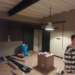 Speedbud uslugi ogolnobudowlane Jakub Nowak - Wnętrze w trakcie remontu: widoczne panele podłogowe, grzejnik elektryczny, lampa wisząca w stylu industrialnym, materiały budowlane i narzędzia, oraz fragment osoby w bluzie z nadrukiem.