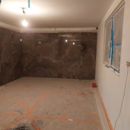 Speedbud uslugi ogolnobudowlane Jakub Nowak - Łazienka w trakcie remontu, ściany wyłożone brązowymi płytami imitującymi marmur, okno zabezpieczone folią, na podłodze widoczne pomarańczowe linie wyznaczające obszar prac.