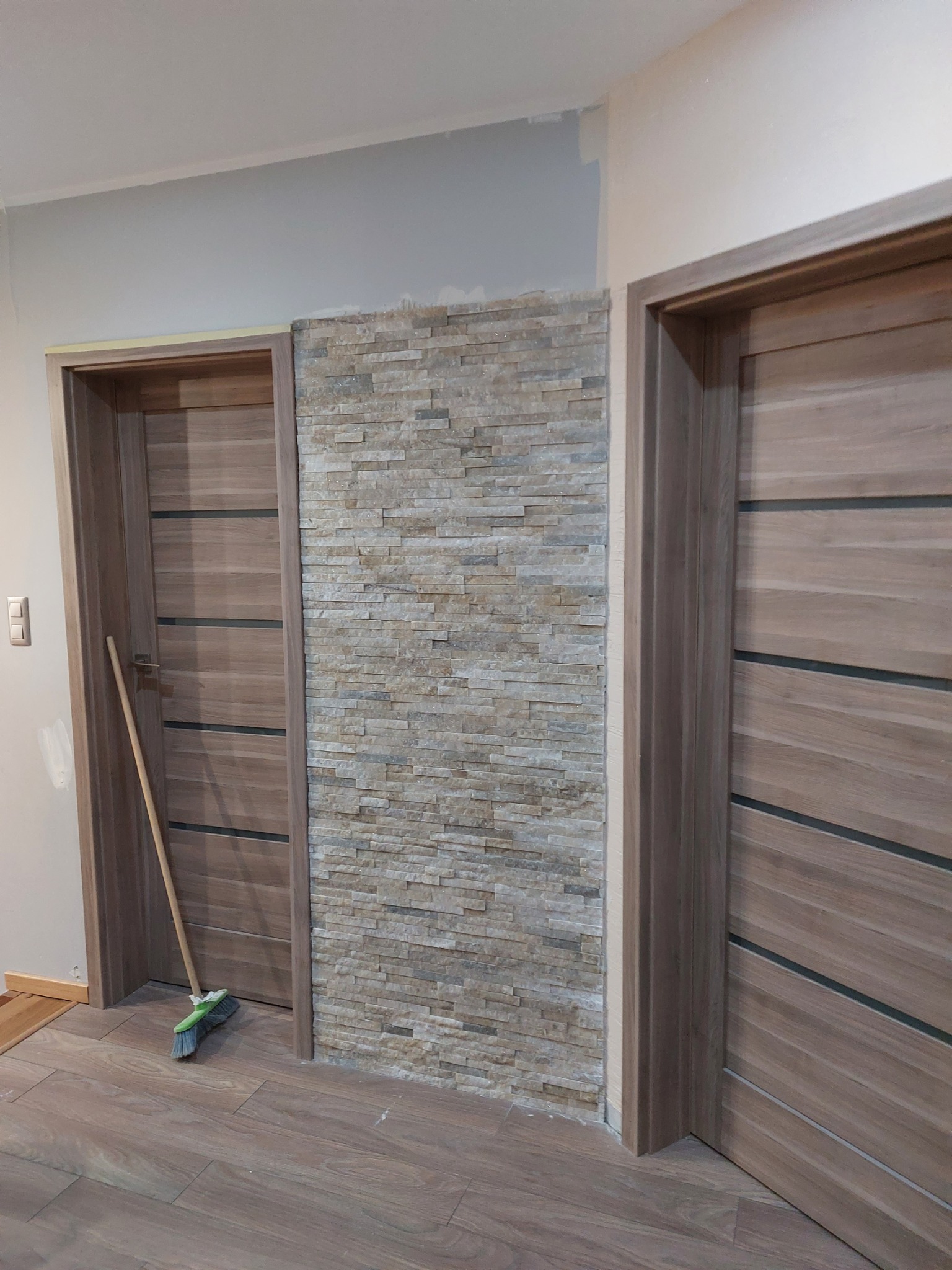Fragment korytarza z wykończoną ścianą z dekoracyjnej cegły, dwiema drewnianymi ościeżnicami drzwiowymi z zamontowanymi drzwiami oraz brudną miotłą opartą o ścianę