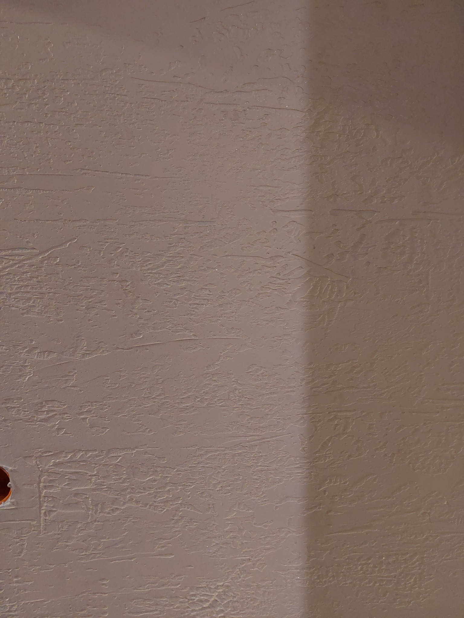 Ściana z wyraźną, poziomą fakturą w odcieniach beżu, z widocznym otworem na gniazdko elektryczne po lewej stronie i delikatnym cieniem po prawej.