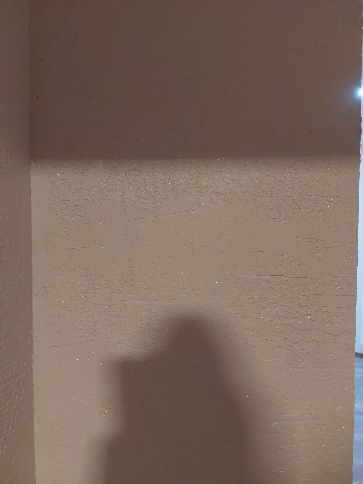 Teksturowana ściana w odcieniach beżu, z widocznym cieniem rzucanym przez obiekt w pomieszczeniu.