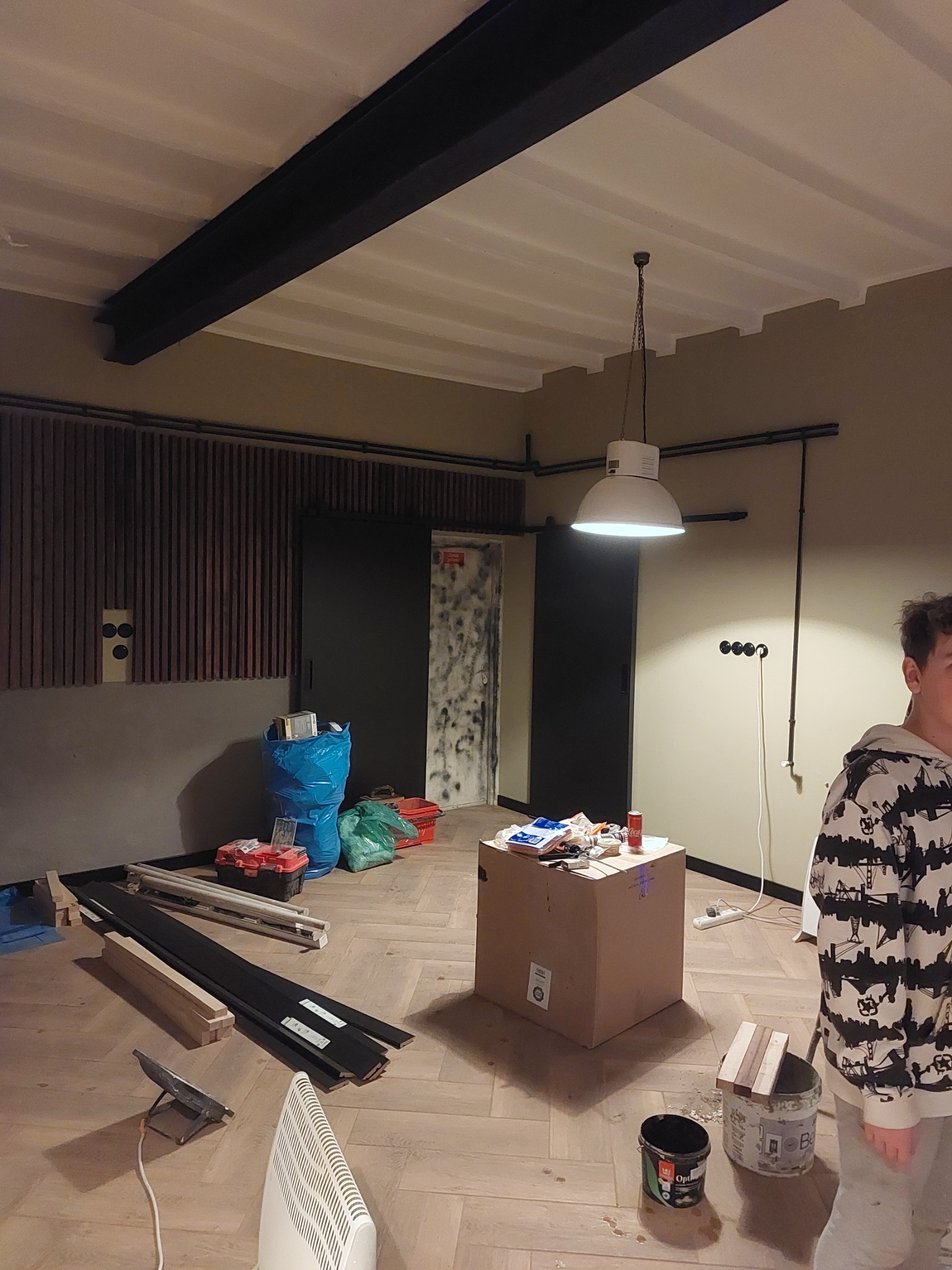 Wnętrze w trakcie remontu: widoczne panele podłogowe, grzejnik elektryczny, lampa wisząca w stylu industrialnym, materiały budowlane i narzędzia, oraz fragment osoby w bluzie z nadrukiem.