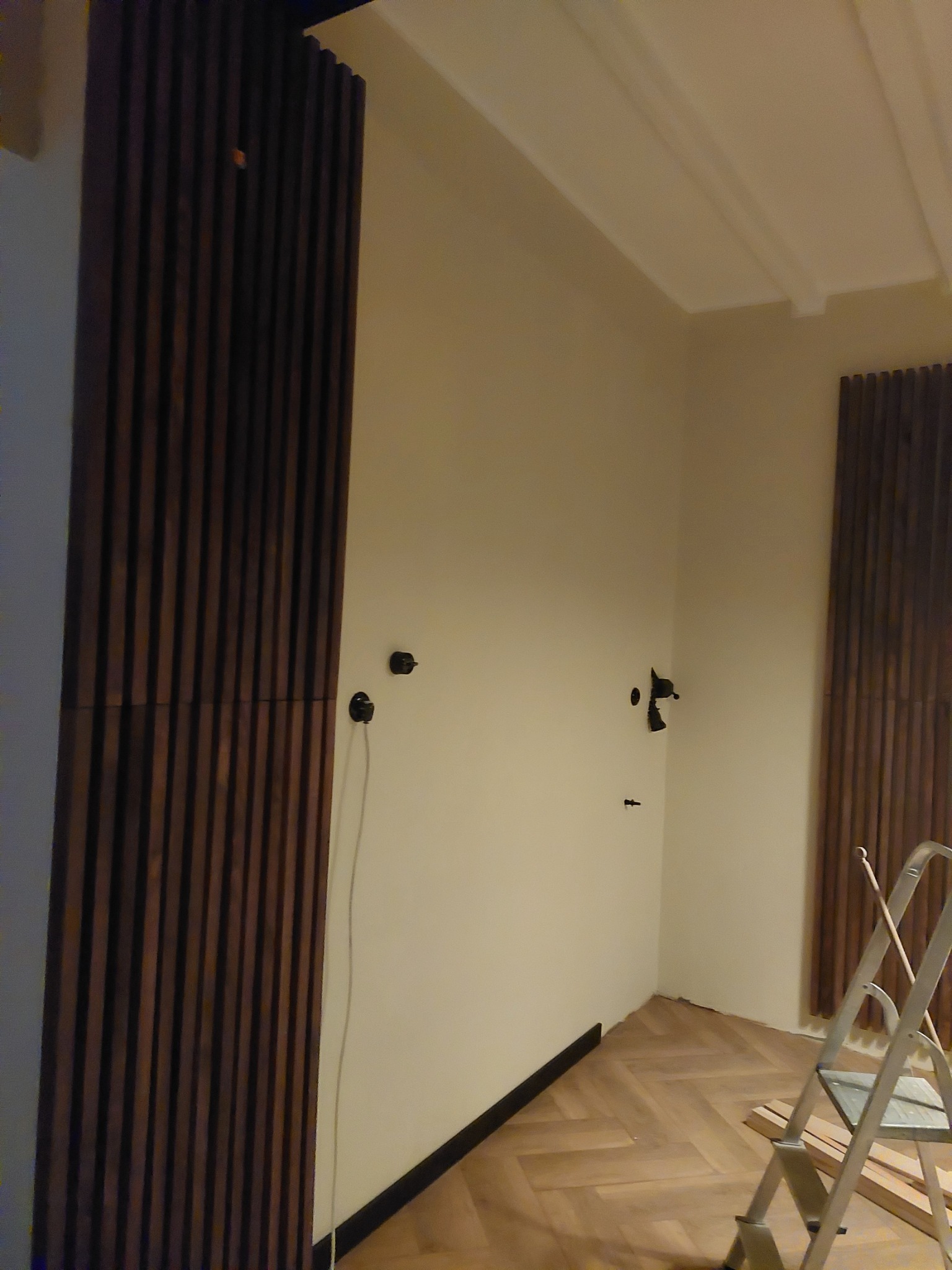 Montaż dekoracyjnych paneli ściennych z ciemnego drewna, widoczne gniazdka elektryczne i przewody, parkiet w jodełkę, drabina malarska w rogu pokoju.