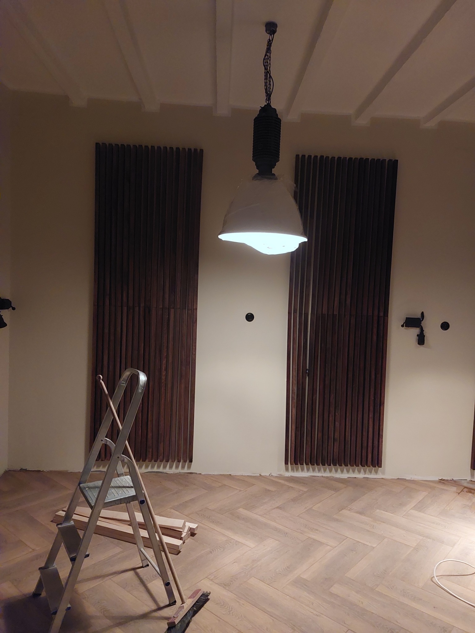 Pokój z drewnianą podłogą ułożoną w jodełkę, na ścianie panele dekoracyjne, drabina aluminiowa i wisząca lampa z kloszem.