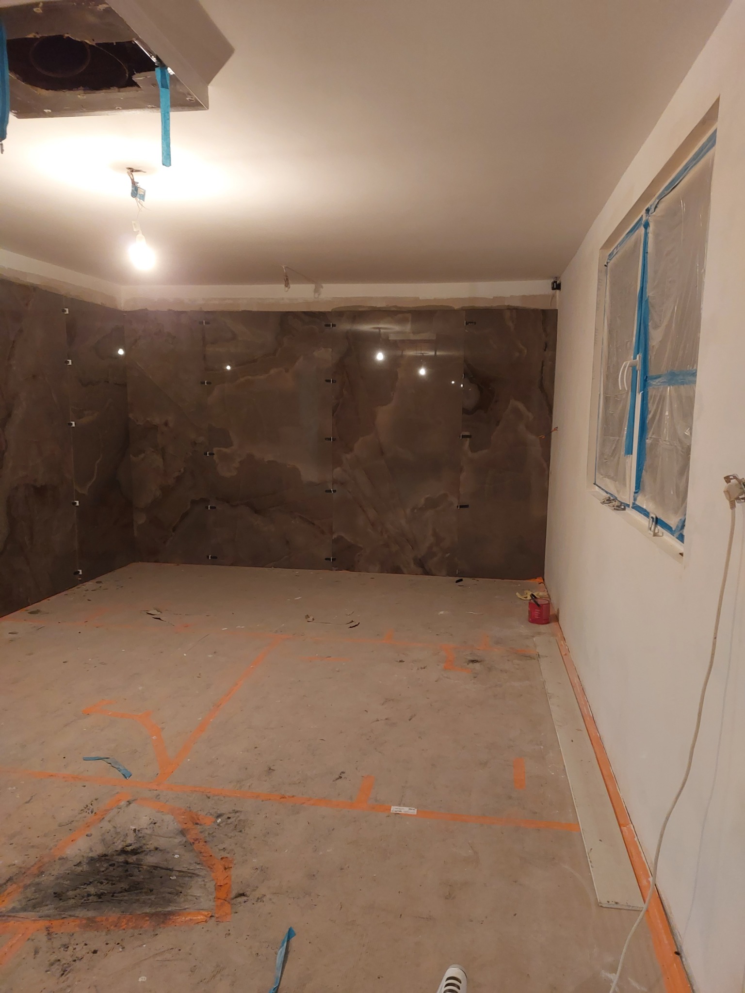 Łazienka w trakcie remontu, ściany wyłożone brązowymi płytami imitującymi marmur, okno zabezpieczone folią, na podłodze widoczne pomarańczowe linie wyznaczające obszar prac.