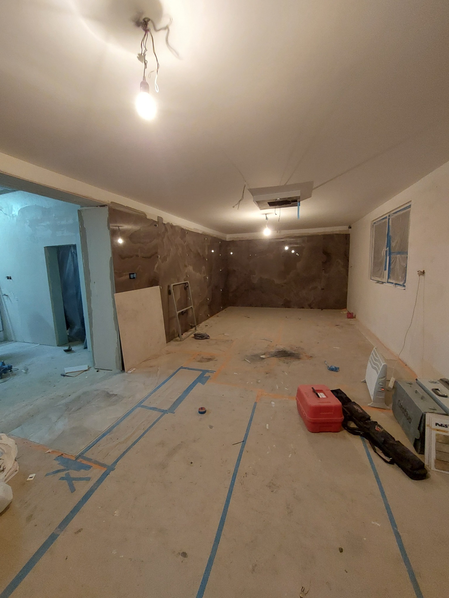 Pokój w trakcie prac wykończeniowych: przygotowane ściany z fakturą, zabezpieczone okno folią, wyznaczona taśmą podłoga, widoczne narzędzia i materiały budowlane.