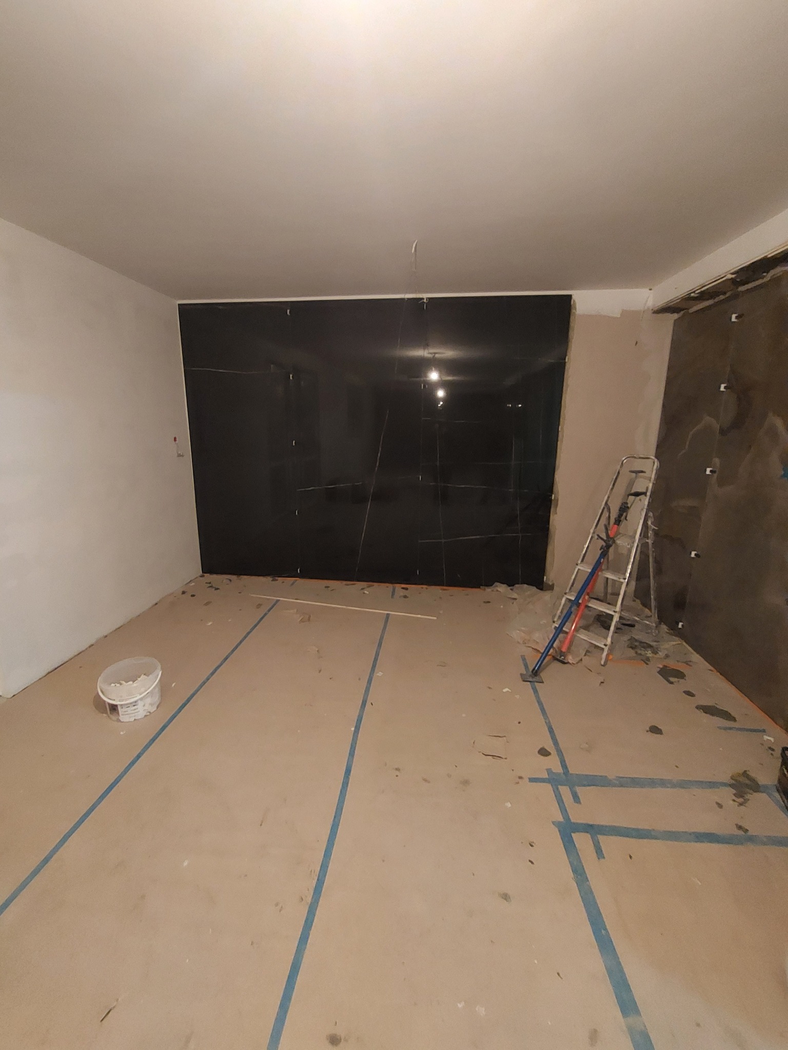 Pokój w trakcie prac wykończeniowych: czarne płytki na ścianie, zabezpieczona podłoga, drabina oparta o ścianę z widocznymi narzędziami.