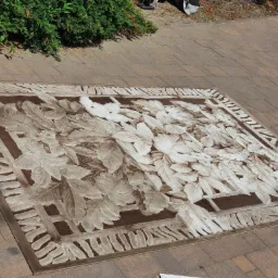 Prostokątny dywan z wzorem liści w odcieniach brązu i beżu leży na kostce brukowej; widoczny krzew w tle i cień osoby.