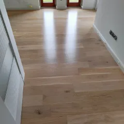 Świeżo położony, lakierowany parkiet dębowy w jasnym odcieniu, widoczne niedokończone detale przy ścianie, niezamaskowane gniazdka elektryczne i przygotowane do malowania ściany.