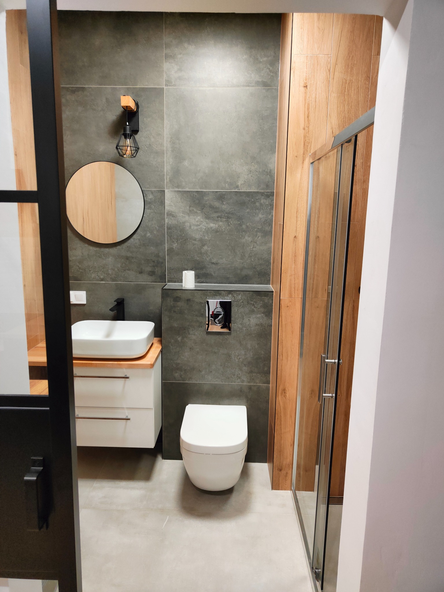 Nowoczesna łazienka z szarymi płytkami, drewnianymi akcentami i czarnymi detalami: okrągłe lustro, designerska lampa i kabina prysznicowa.