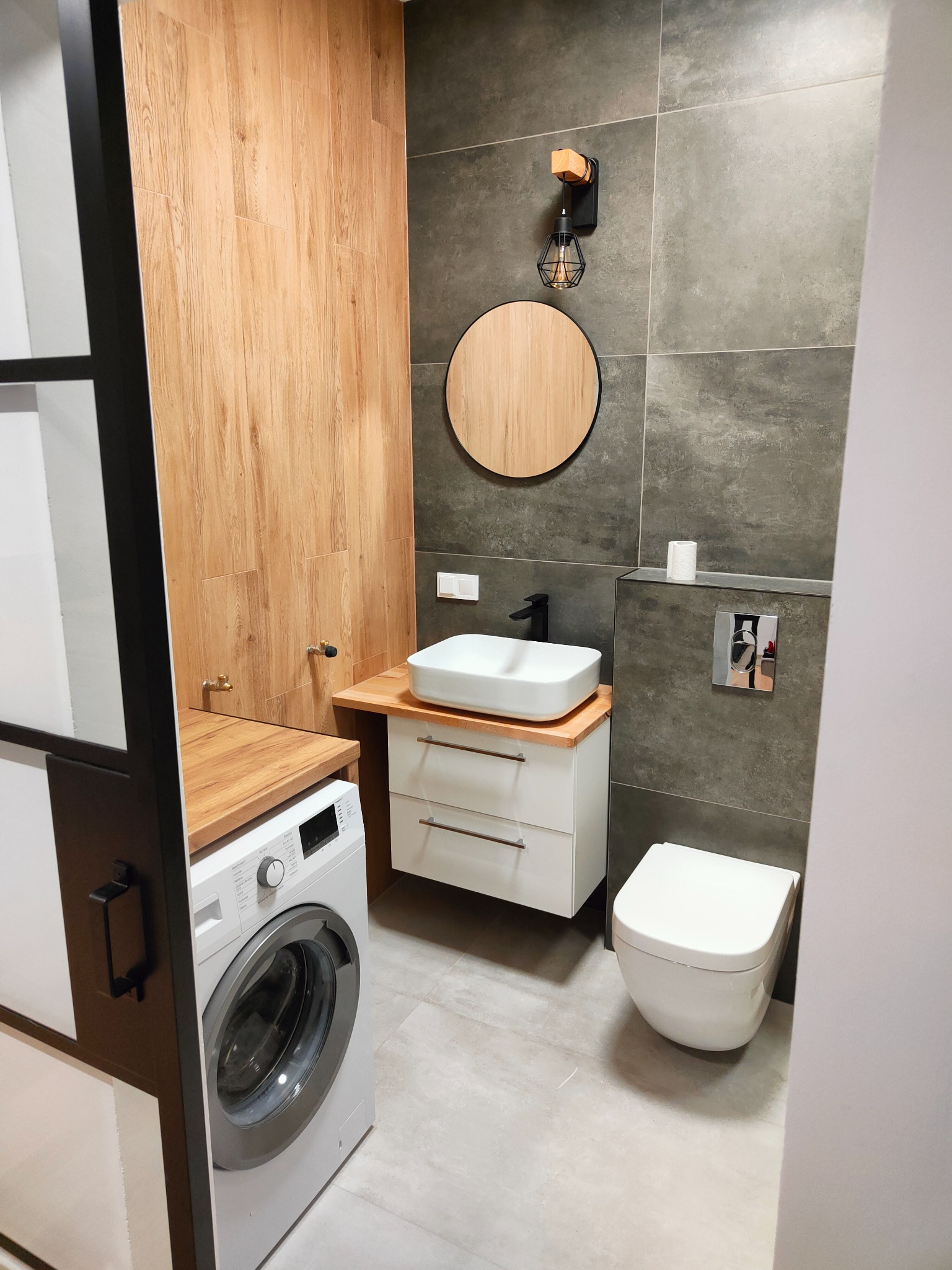 Nowoczesna łazienka z drewnianymi akcentami: umywalka nablatowa, okrągłe lustro, szara glazura, pralka i toaleta podwieszana.
