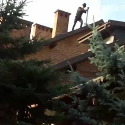 Kominiarz w Lesku w trakcie pracy na dachu ceglanego domu, widoczny komin i narzędzie do czyszczenia.