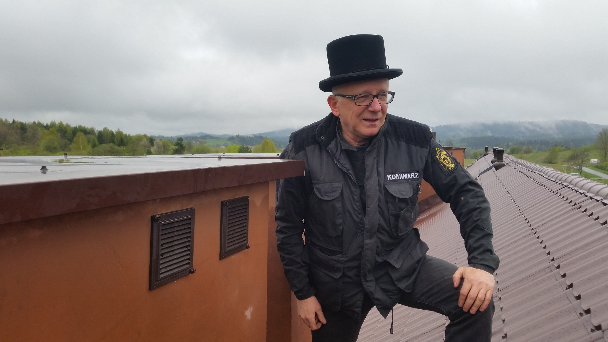 Kominiarz w czarnym cylindrze i kurtce z naszywką 'KOMINIARZ' stoi na mokrym, brązowym dachu, z widokiem na zalesione wzgórza w tle.