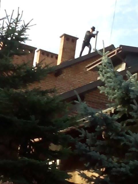 Kominiarz w Lesku w trakcie pracy na dachu ceglanego domu, widoczny komin i narzędzie do czyszczenia.