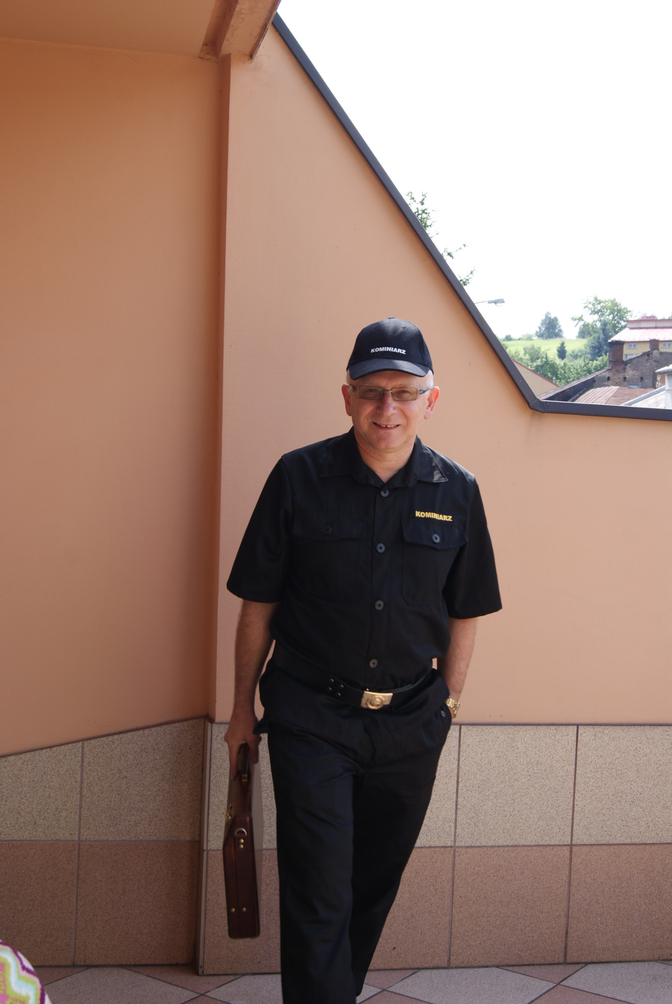 Uśmiechnięty mężczyzna w czarnym uniformie z napisem 'KOMINIARZ' na czapce i koszuli, trzymający brązową aktówkę, stoi na zewnątrz budynku z beżową ścianą i kafelkowym cokołem.