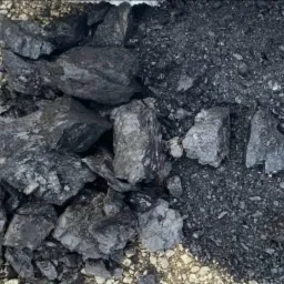 Góra brył węgla kamiennego, częściowo rozdrobnionego, ułożona na ziemi z widocznymi drobnymi kamykami.