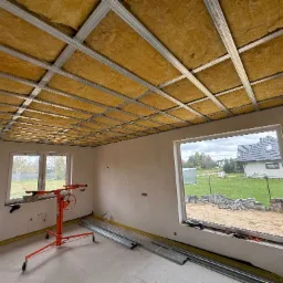 Montaż izolacji z wełny mineralnej w metalowym stelażu sufitu podwieszanego w nowym domu, widoczny podnośnik do płyt oraz profile, okna z widokiem na posesję.