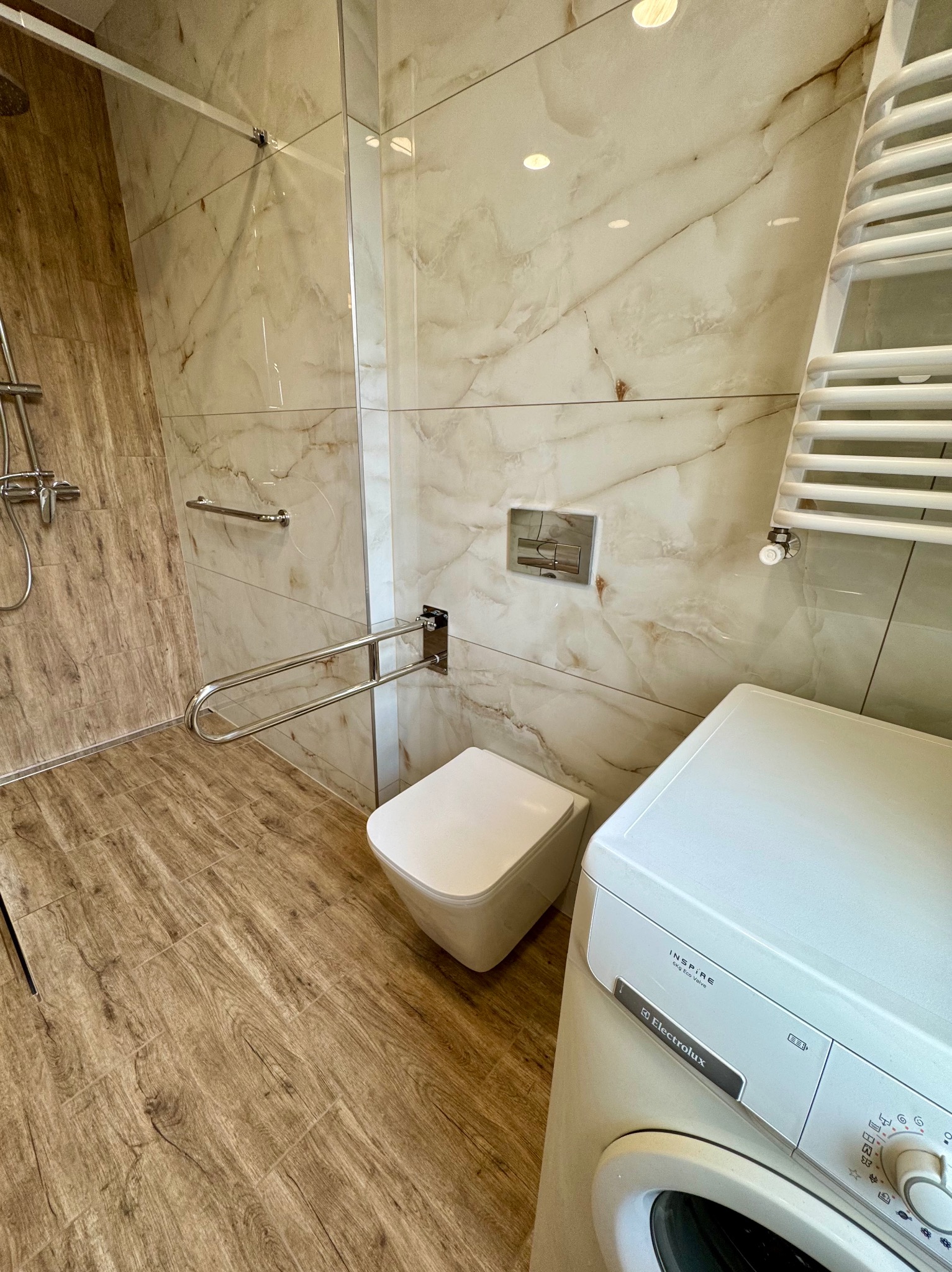 Wykończona łazienka z beżowymi płytkami imitującymi marmur, drewnopodobną podłogą, toaletą, pralką i uchwytami dla niepełnosprawnych. Nowoczesny design.