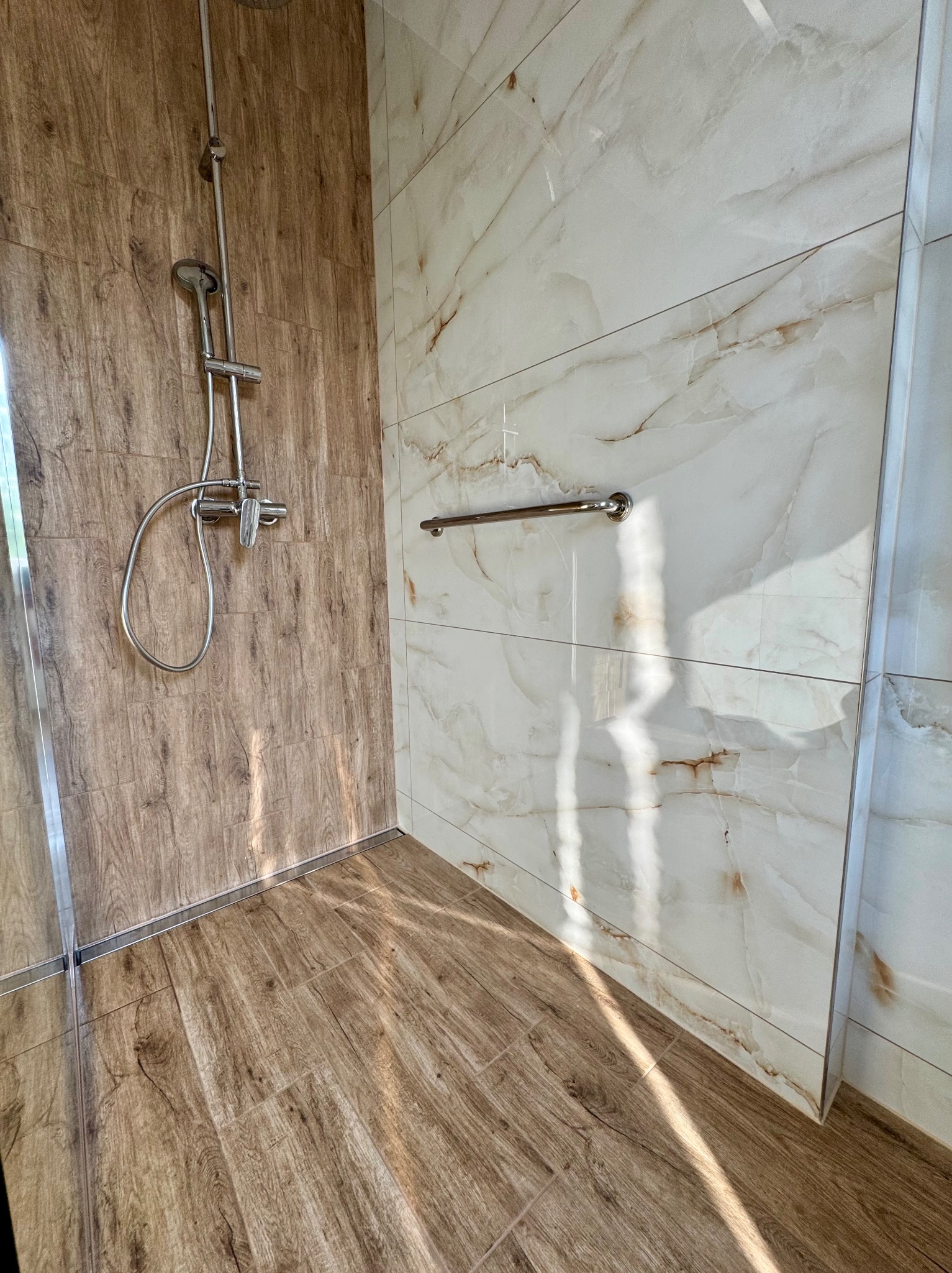 Nowoczesna kabina prysznicowa z drewnopodobnymi i marmurowymi płytkami, chromowaną armaturą i uchwytem, eleganckie wykończenie.