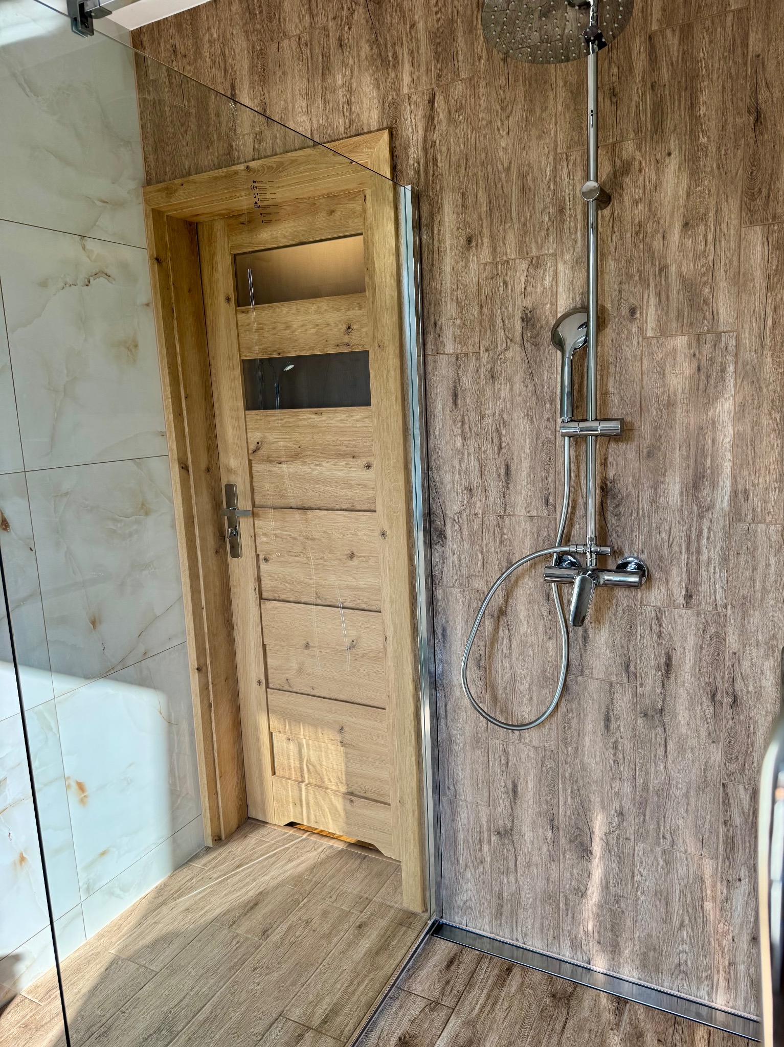 Wykończona łazienka: drewniane drzwi z szybami, chromowana armatura prysznicowa, marmurowe i drewnopodobne płytki na ścianach, szklana kabina.