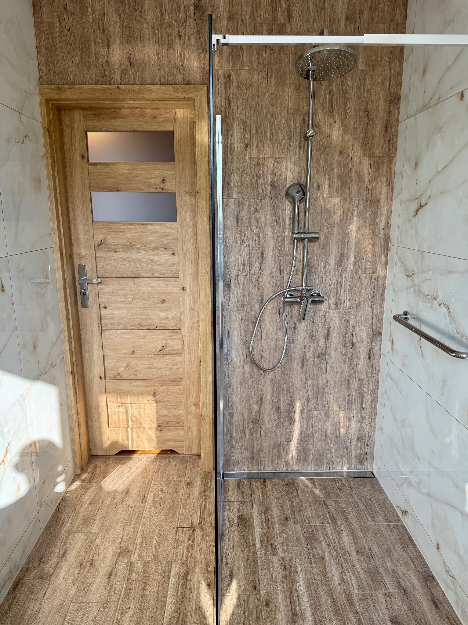 Nowoczesna łazienka z kabiną typu walk-in, drewnopodobnymi płytkami i drewnianymi drzwiami z szybą. Bateria prysznicowa w kolorze chromu.