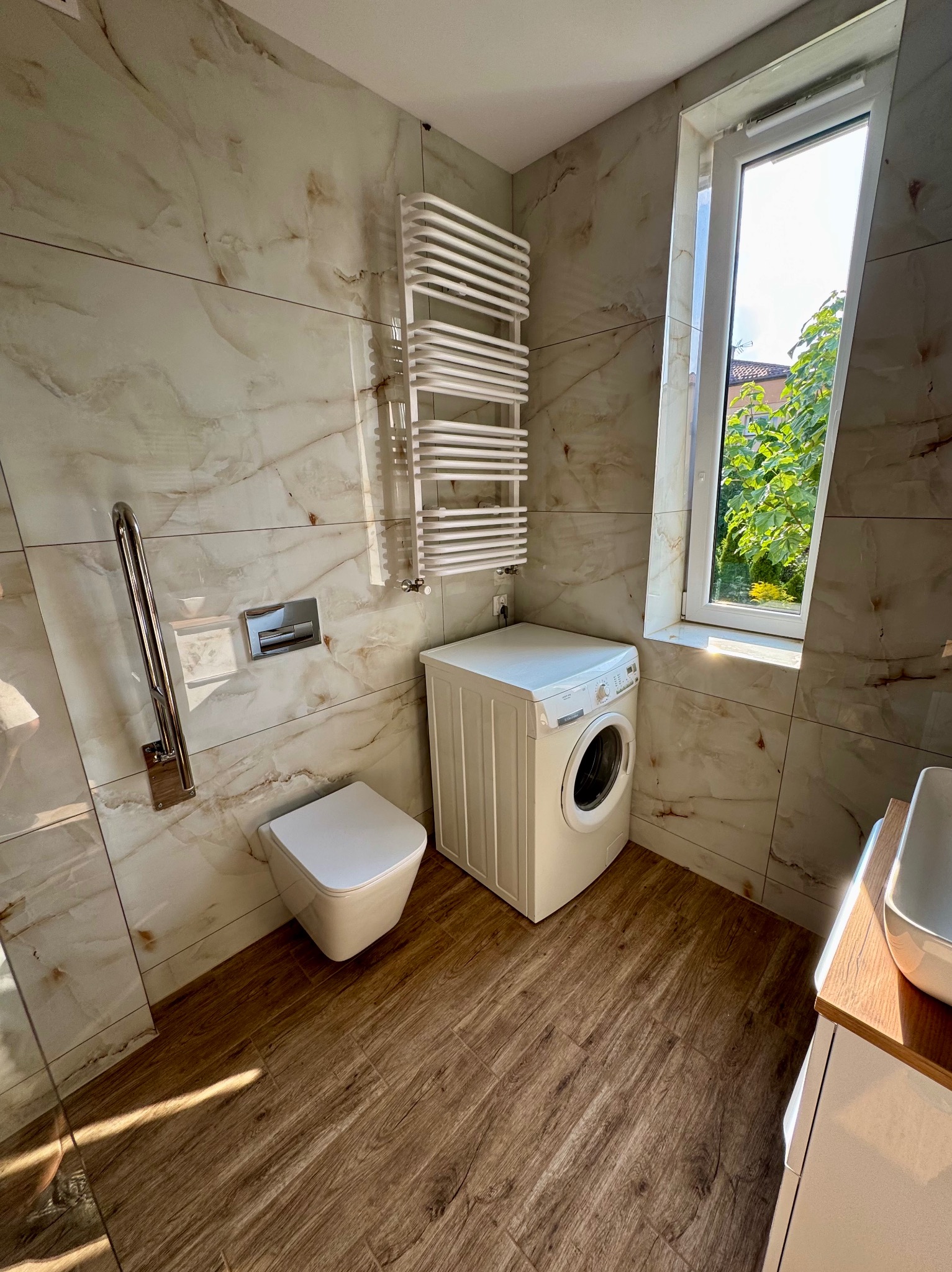 Jasna łazienka z marmurowymi płytkami, drewnopodobną podłogą i białym wyposażeniem: toaleta, pralka, grzejnik drabinkowy i reling dla niepełnosprawnych.