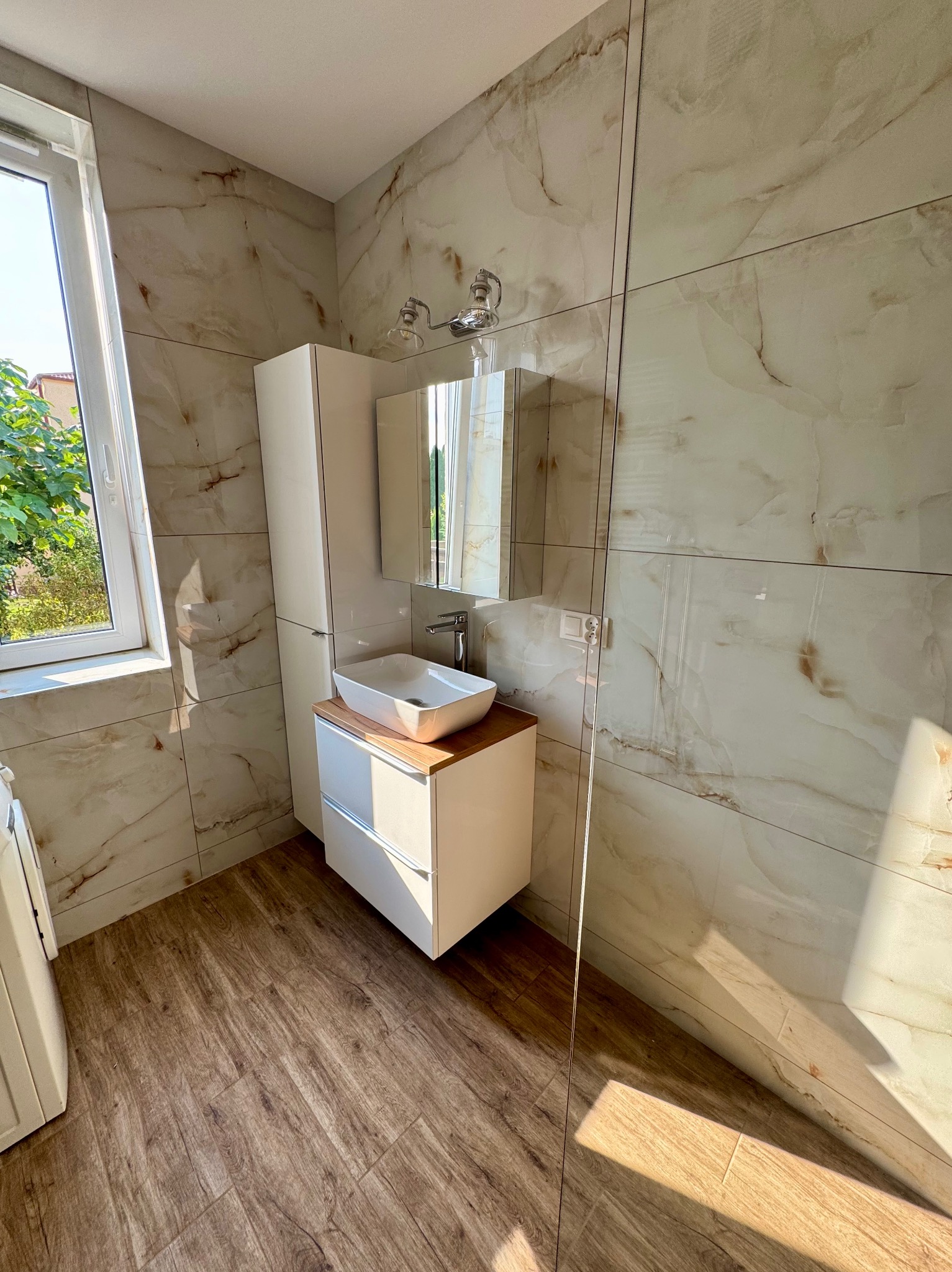 Wykończona łazienka z marmurowymi płytkami ściennymi, drewnopodobną podłogą i białymi szafkami. Umywalka nablatowa i lustro z oświetleniem.