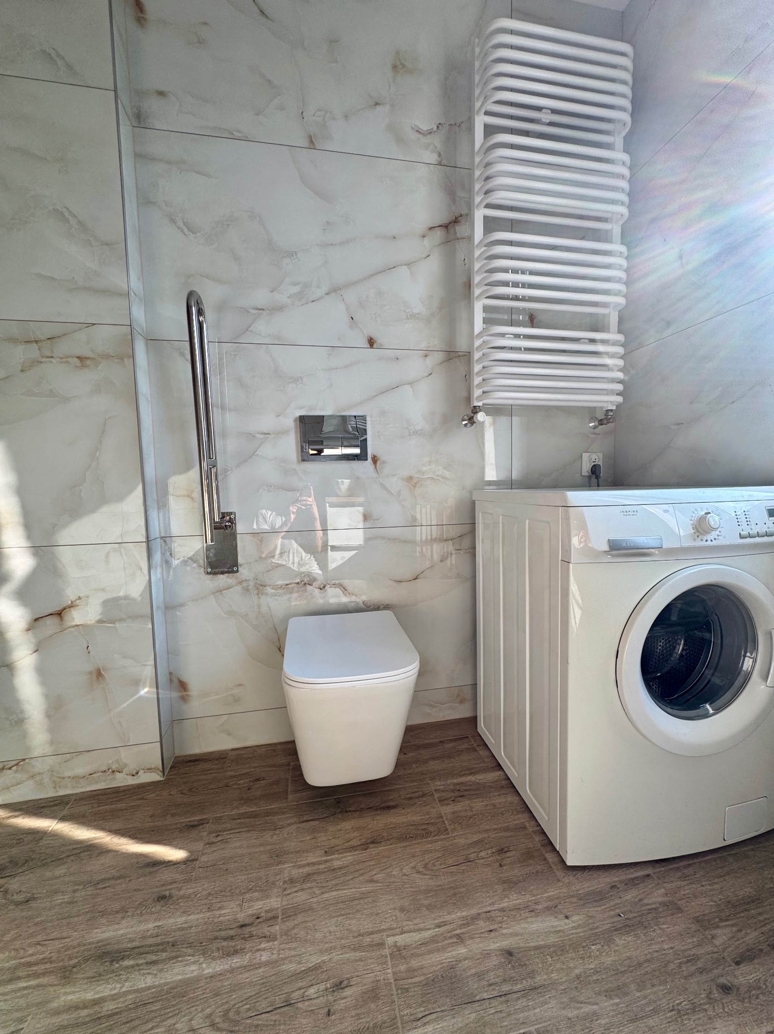 Wykończona łazienka z marmurowymi płytkami, drewnopodobną podłogą, grzejnikiem drabinkowym i pralką. Uchwyt dla niepełnosprawnych obok toalety.