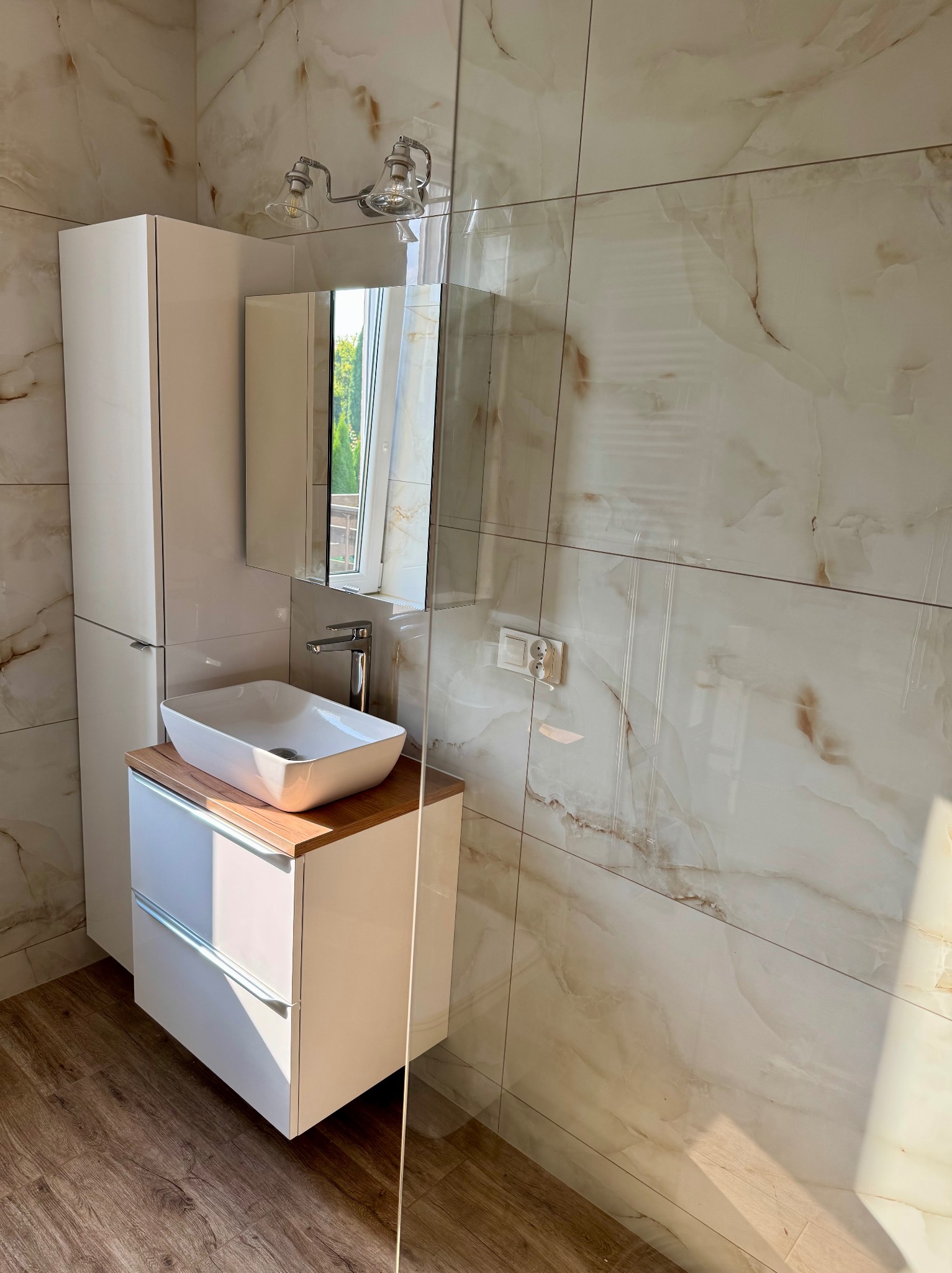 Biała umywalka nablatowa na drewnianym blacie, szafka z szufladami, lustro i oświetlenie. Płytki imitujące marmur na ścianach, drewnopodobna podłoga.