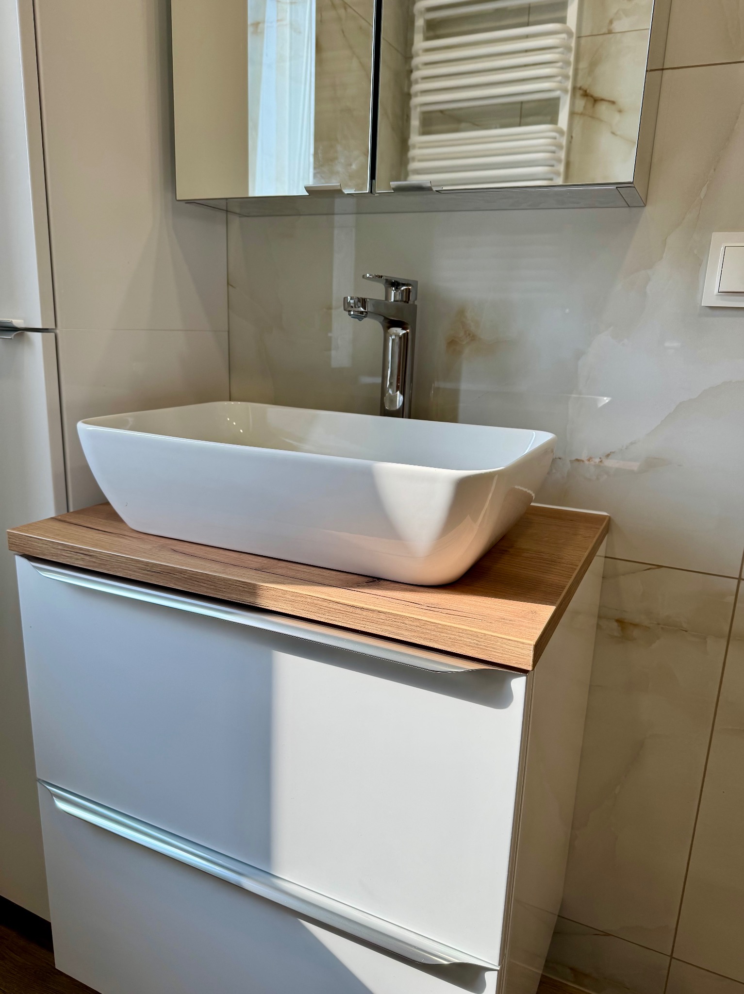 Biała, prostokątna umywalka nablatowa na szafce z drewnianym blatem w łazience wykończonej płytkami imitującymi marmur. Bateria chromowana, lustro z szafką.