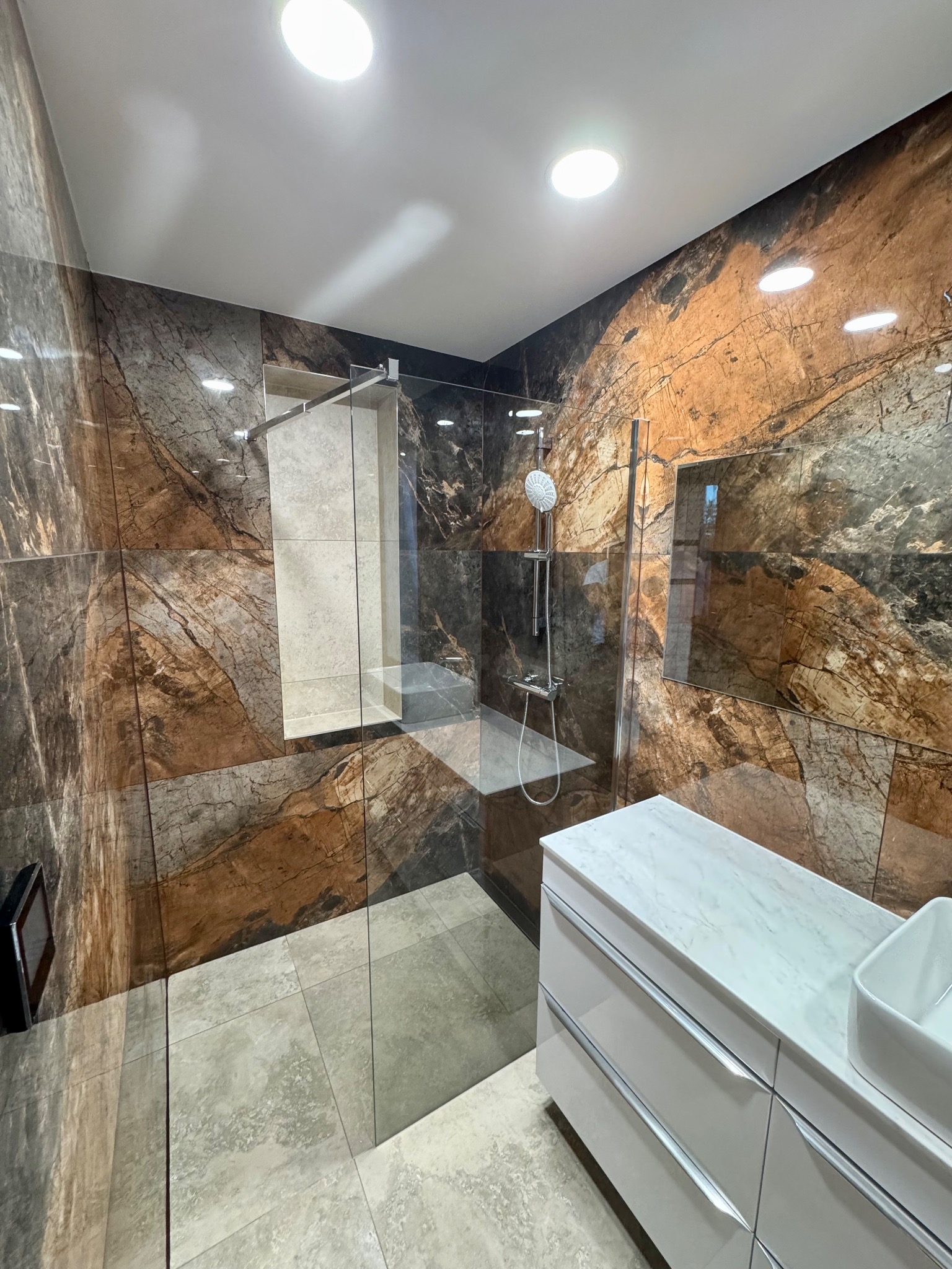 Nowoczesna łazienka z kabiną typu walk-in, marmurowe płytki na ścianach i podłodze, biała szafka z umywalką. Widoczne oświetlenie punktowe.