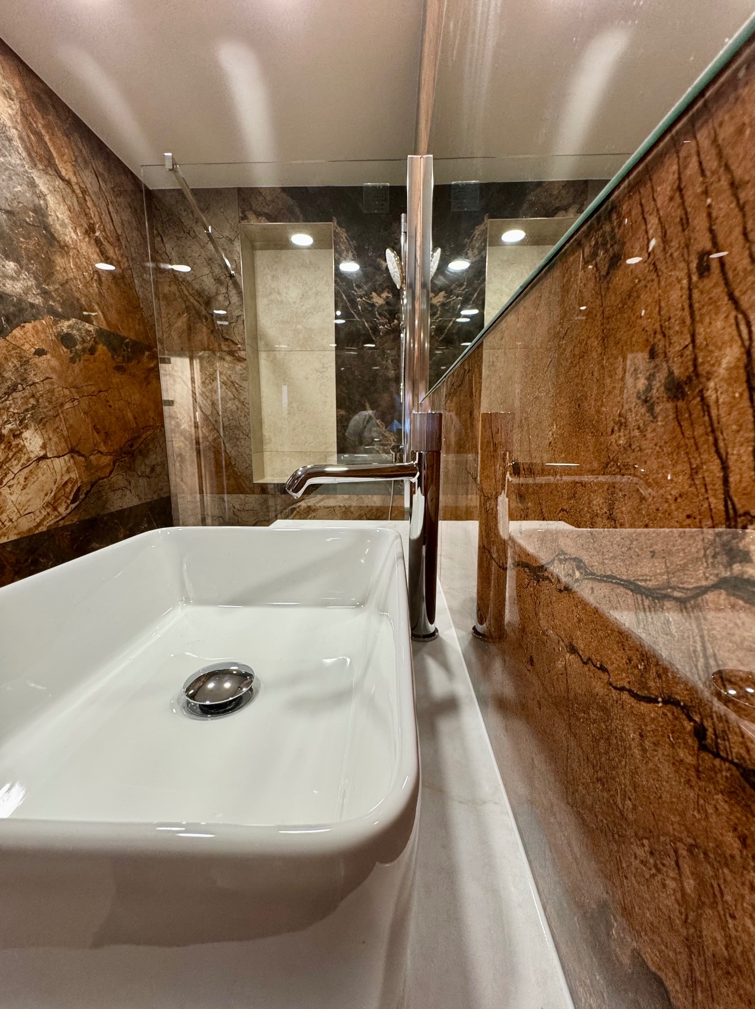 Biała, prostokątna umywalka z chromowaną baterią na tle ściany z brązowego marmuru w luksusowej łazience. Widoczne odbicie w lustrze.