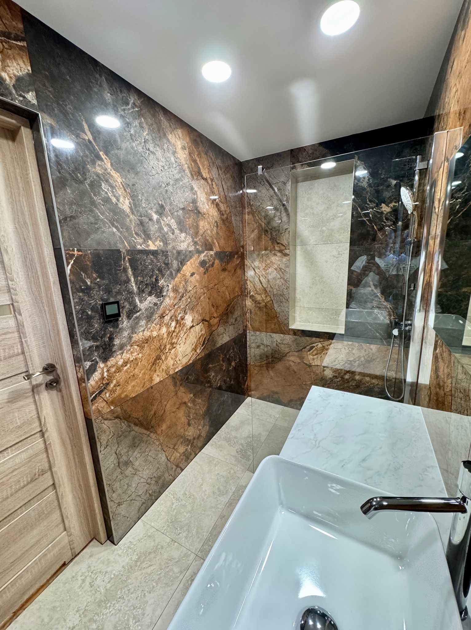 Elegancka łazienka po remoncie: kabina prysznicowa ze szklaną ścianą, marmurowe płytki na ścianach i podłodze, nowoczesna umywalka nablatowa, drewniane drzwi.