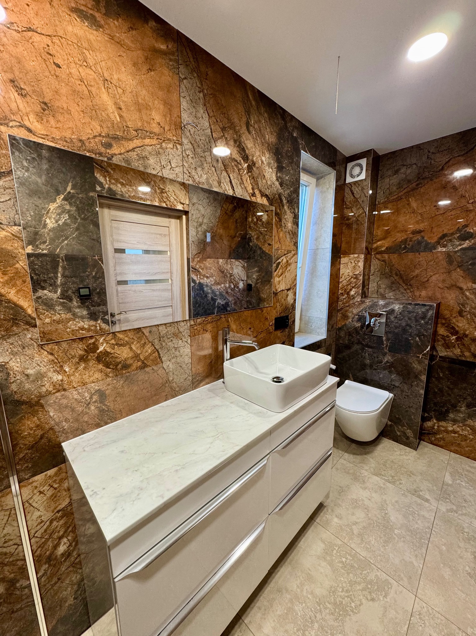 Elegancka łazienka z marmurowymi płytkami, białą umywalką nablatową i toaletą podwieszaną. W lustrze odbicie drzwi. Lampa sufitowa. Wykończenie w ciepłych brązach.