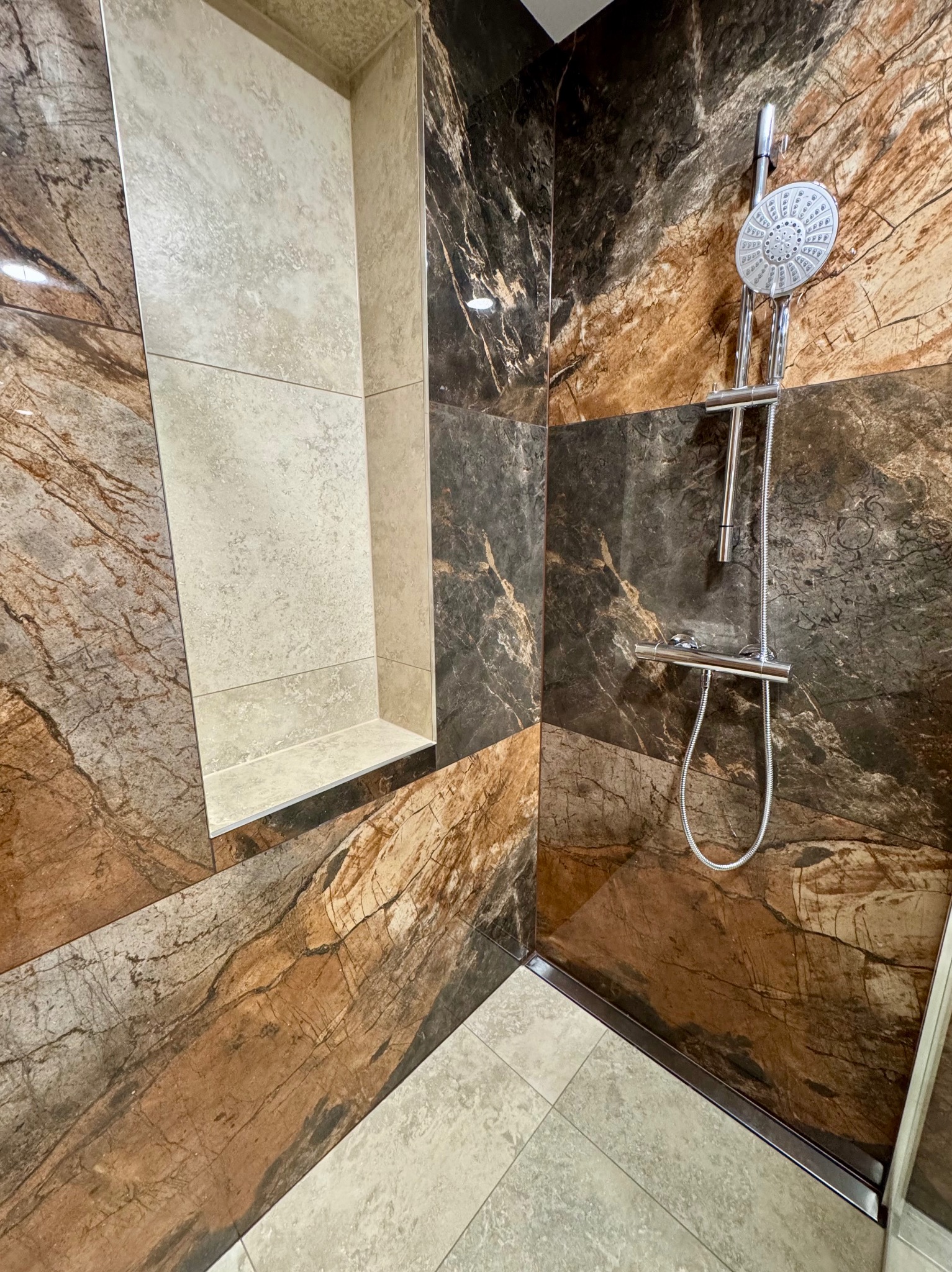 Nowoczesna kabina prysznicowa z marmurowymi płytkami w odcieniach brązu i beżu, z wnęką wyłożoną jasnymi płytkami i chromowaną armaturą.