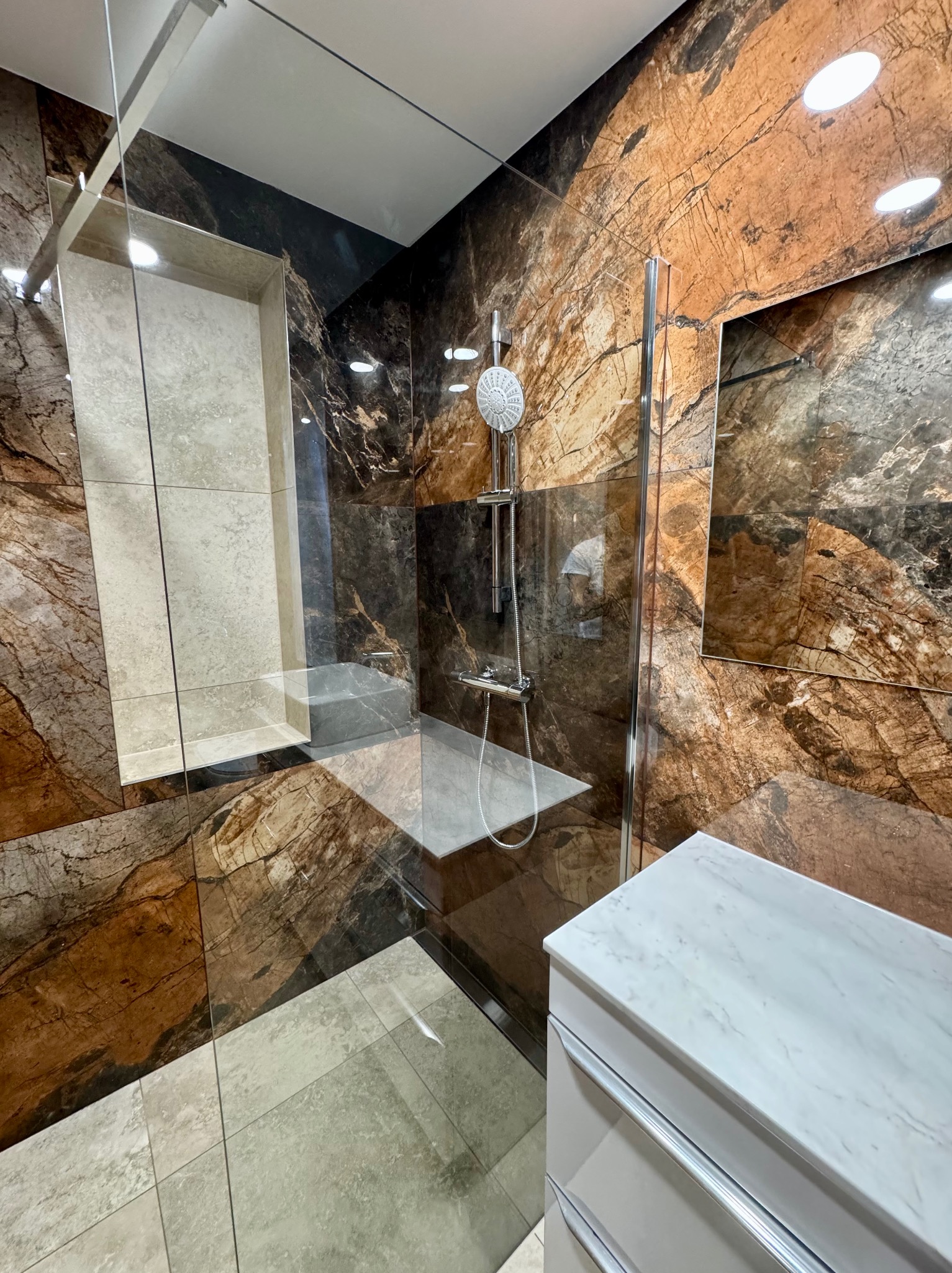Nowoczesna kabina prysznicowa z marmurowymi płytkami w odcieniach brązu i szarości, przeszklona ściana, szafka z marmurowym blatem. Efektowny design łazienki.