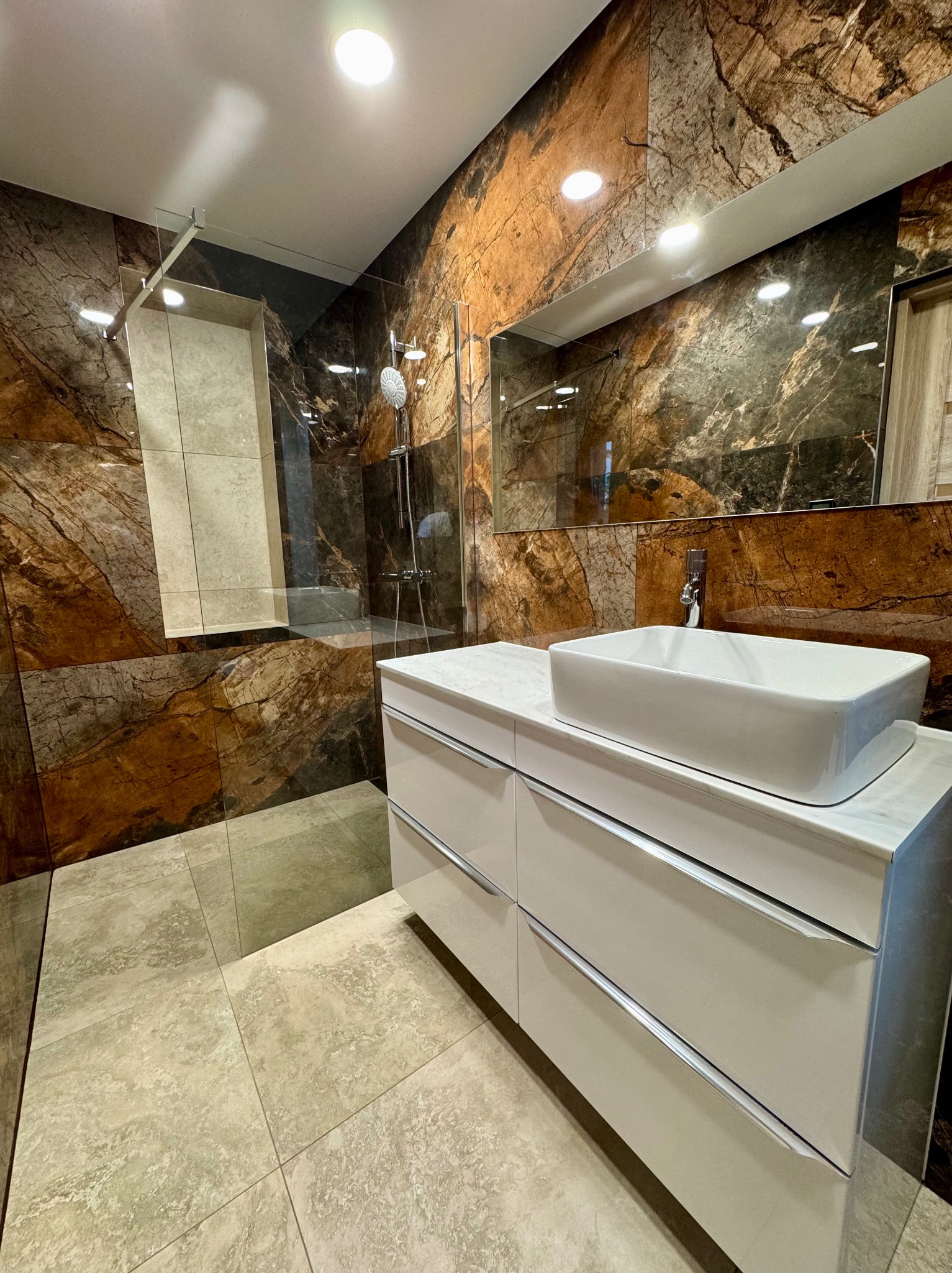 Elegancka łazienka z kabiną prysznicową typu walk-in, umywalką nablatową na szafce z szufladami i lustrem. Ściany wykończone płytkami imitującymi marmur w ciepłych odcieniach.