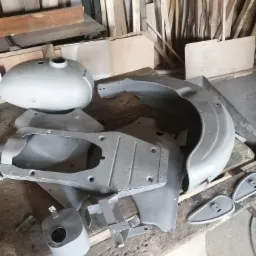 Elementy motocykla po obróbce przygotowane do dalszych prac, widoczne: zbiornik paliwa, błotnik, podnóżki i inne drobne części, tło stanowią deski i drewniane elementy.