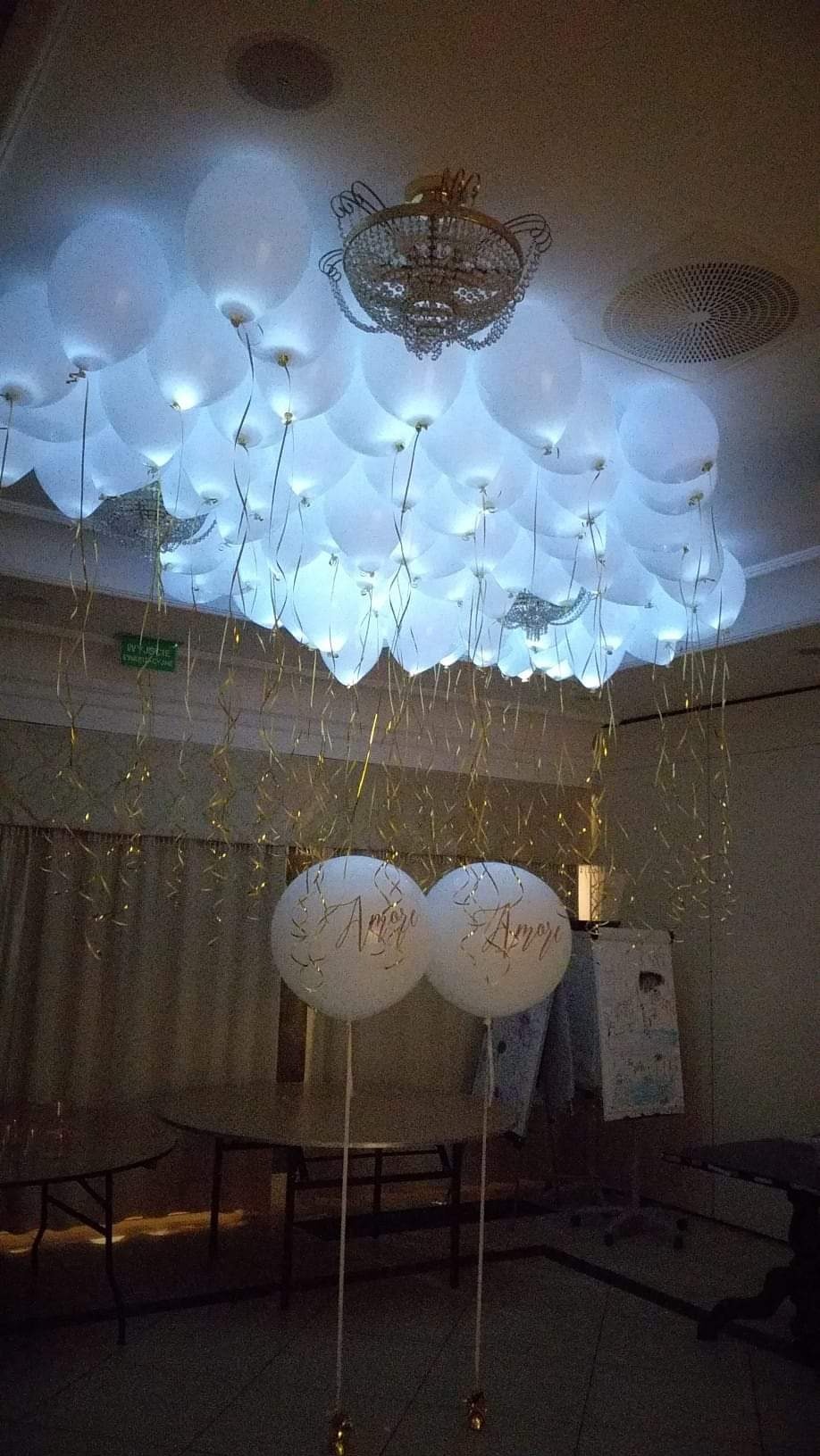 Dekoracja sali balowej: białe, podświetlane balony pod sufitem, zwisające złote wstążki, dwa balony z napisem 'Amori' na pierwszym planie, żyrandol w tle.