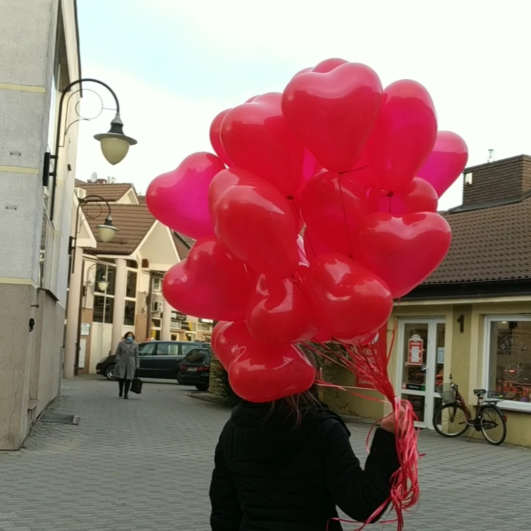 Osoba trzyma bukiet czerwonych balonów w kształcie serc na ulicy, w tle budynki i przechodząca osoba w płaszczu i maseczce.