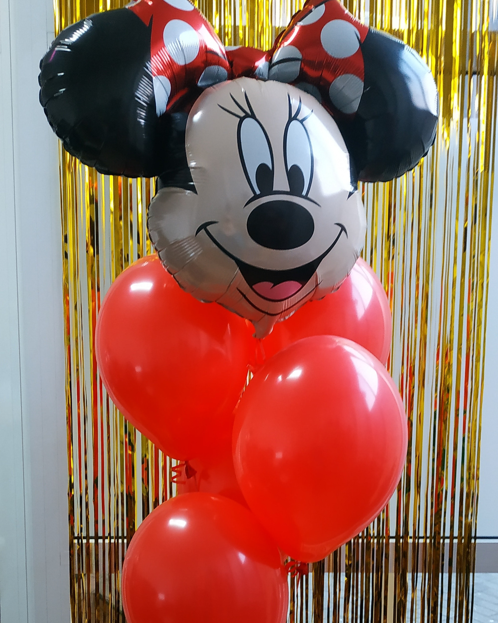 Balon foliowy Myszki Minnie z czerwoną kokardą w białe kropki, otoczony czerwonymi balonami lateksowymi na tle złotych frędzli.