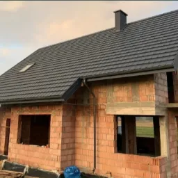 Dom w stanie surowym otwartym w Lublinie z nowym, grafitowym pokryciem dachowym z blachodachówki i orynnowaniem. Widoczny komin i okno dachowe.