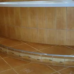 Brązowe płytki ceramiczne ułożone na podłodze i ścianie w łazience, tworzące obudowę wanny z charakterystycznym, ozdobnym pasem płytek na krawędzi stopnia.