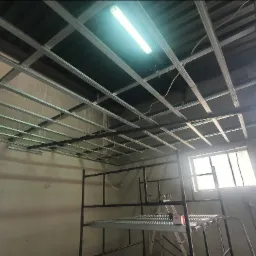 Montaż metalowej konstrukcji sufitu podwieszanego z płyty karton gips, widoczne rusztowanie i okno w surowym wnętrzu.