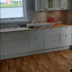 Montaż szarych szafek kuchennych z przeszklonymi górnymi drzwiczkami i złotymi gałkami, widoczne elementy instalacji elektrycznej i przygotowane panele podłogowe.