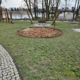 Okrągła rabata z korą i sadzonkami na trawniku w otoczeniu placu zabaw z huśtawką oponą i widokiem na rzekę w tle.